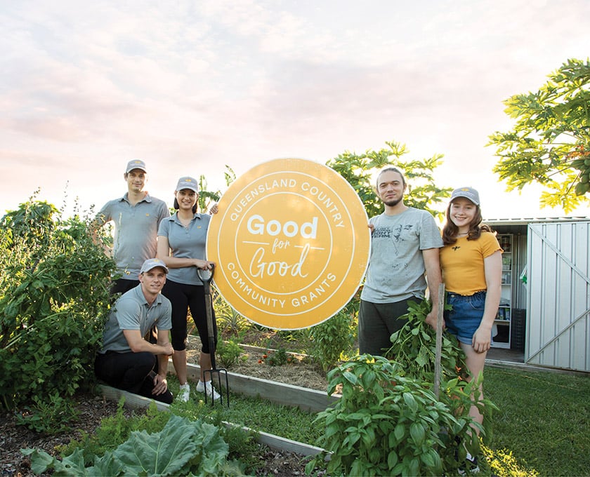 A group of staff stand in a community garden holding a "Good for Good Community Grants" sign